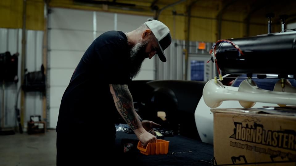 Person working on a car engine in a dim garage, bent over with tools beside a white vehicle.