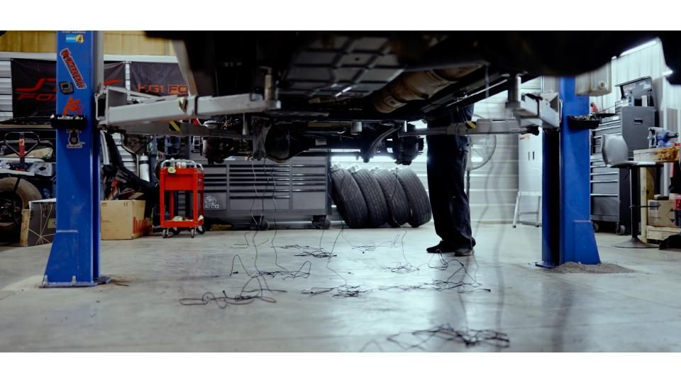 Low-angle view of a car in a mechanic’s garage on a lift, with tools and a mechanic visible underneath.