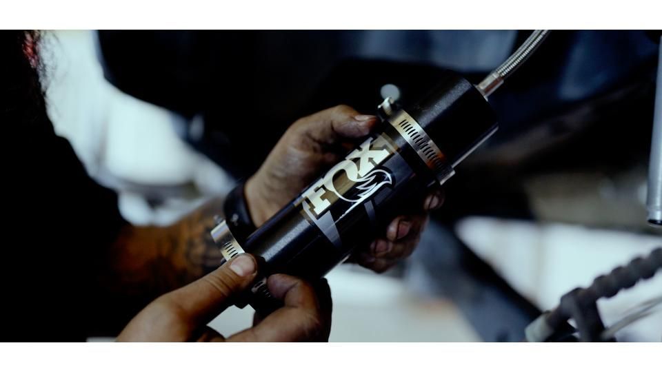 Hands holding a dark cylindrical mechanical part with a metal fitting and wires in a workshop setting