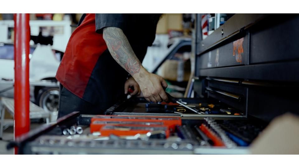 Mechanic working on an open tool chest beside a red vehicle in a garage