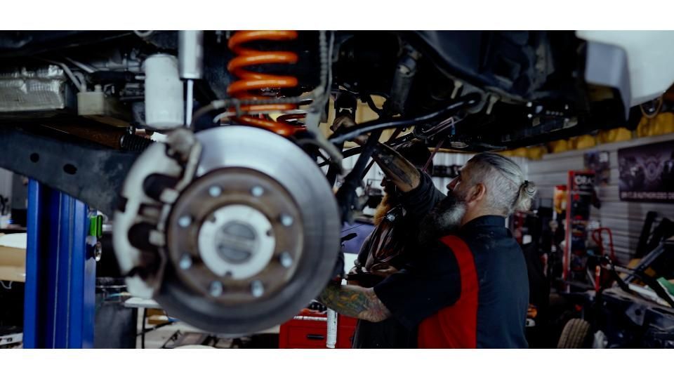 Close-up of a car wheel and suspension in a garage, with a mechanic working underneath.