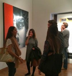 Gente charlando en una galería de arte junto a un gran retrato en blanco y negro.