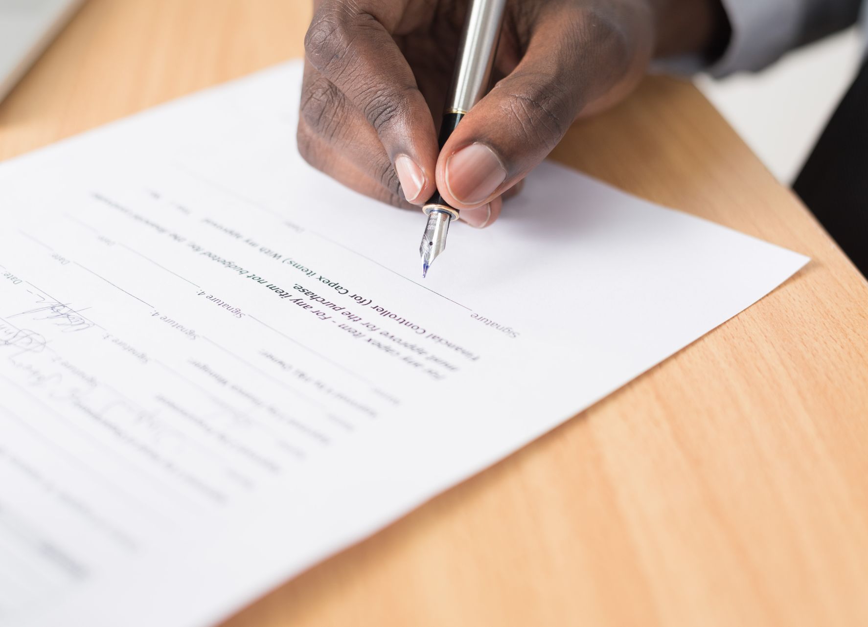 Black hand signing a document with a pen on a light wood desk.