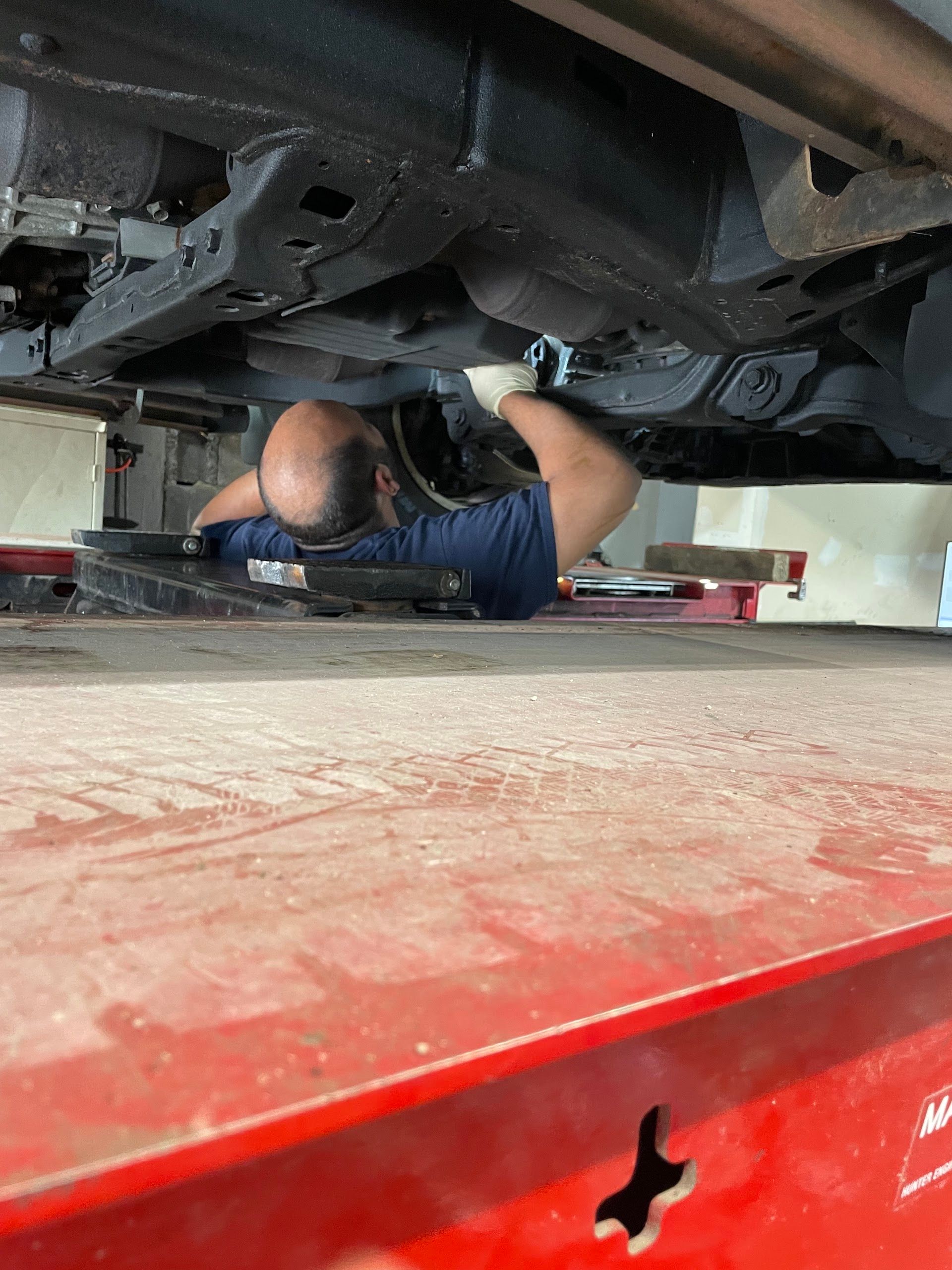 A mechanic lying under a vehicle, working on the undercarriage. Red lift, dark metal, and wearing gloves. | A & R Auto Repair