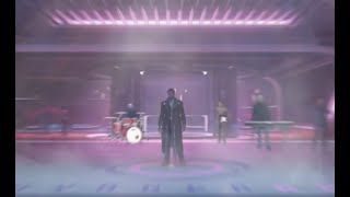 A band performs on a stage in a dimly lit venue with purple hues. A singer stands center stage with a drummer, keyboardist, and guitarist in the background.