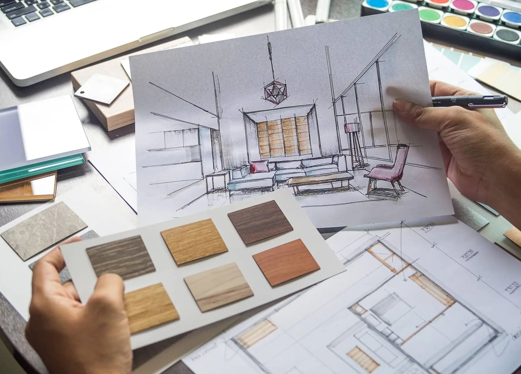 Hand holding an interior design sketch with material samples and floor plans on a desk