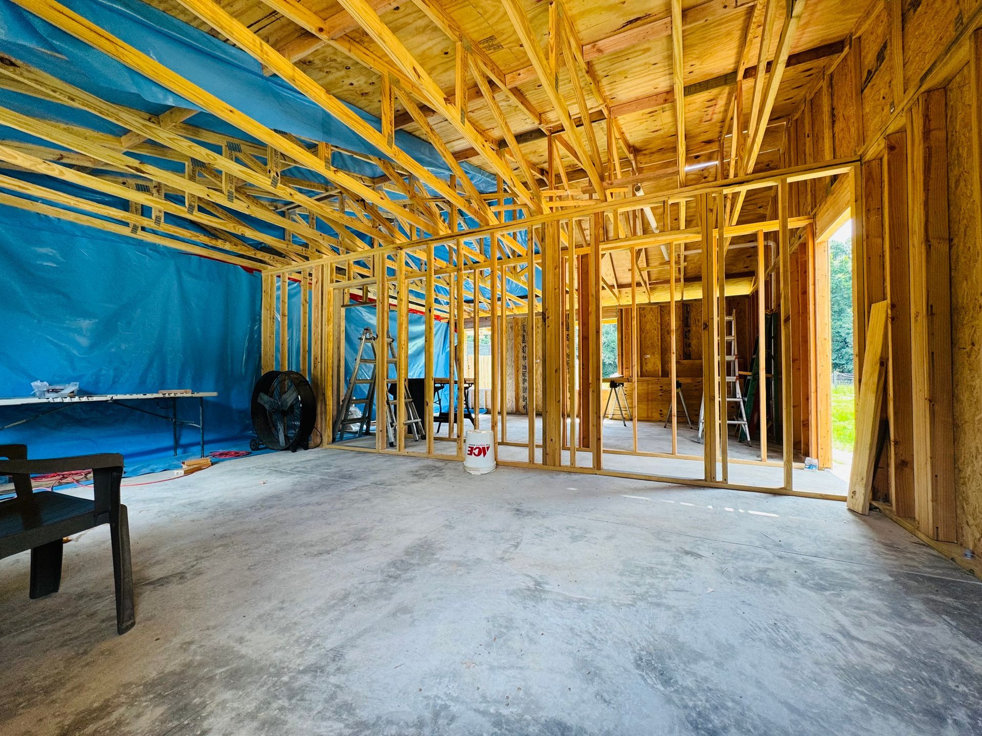 Framed house interior under construction with exposed wood studs, blue tarp wall, and unfinished concrete floor