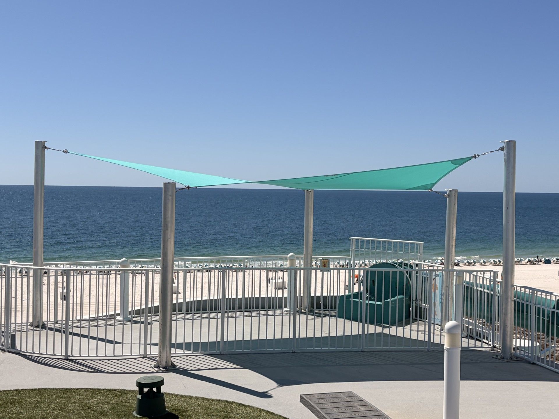 Beachside shaded seating area with turquoise canopy, railing, and ocean view under clear blue sky