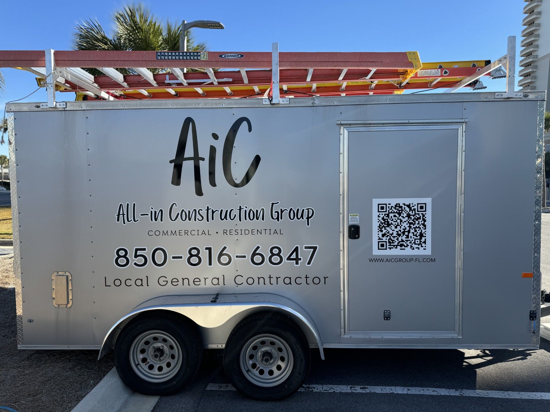 Silver utility trailer with AIC logo, phone number, QR code, and roof-mounted equipment against a blue sky
