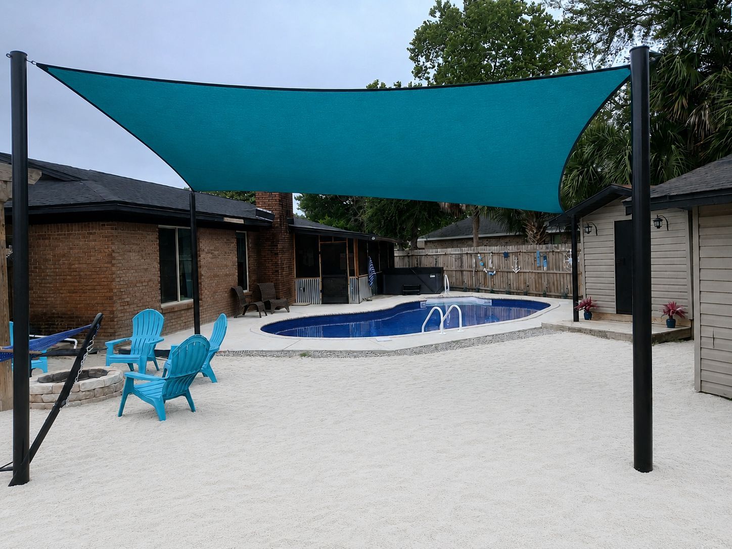 Backyard pool with teal shade sail, blue chairs, and a brick patio under overcast sky
