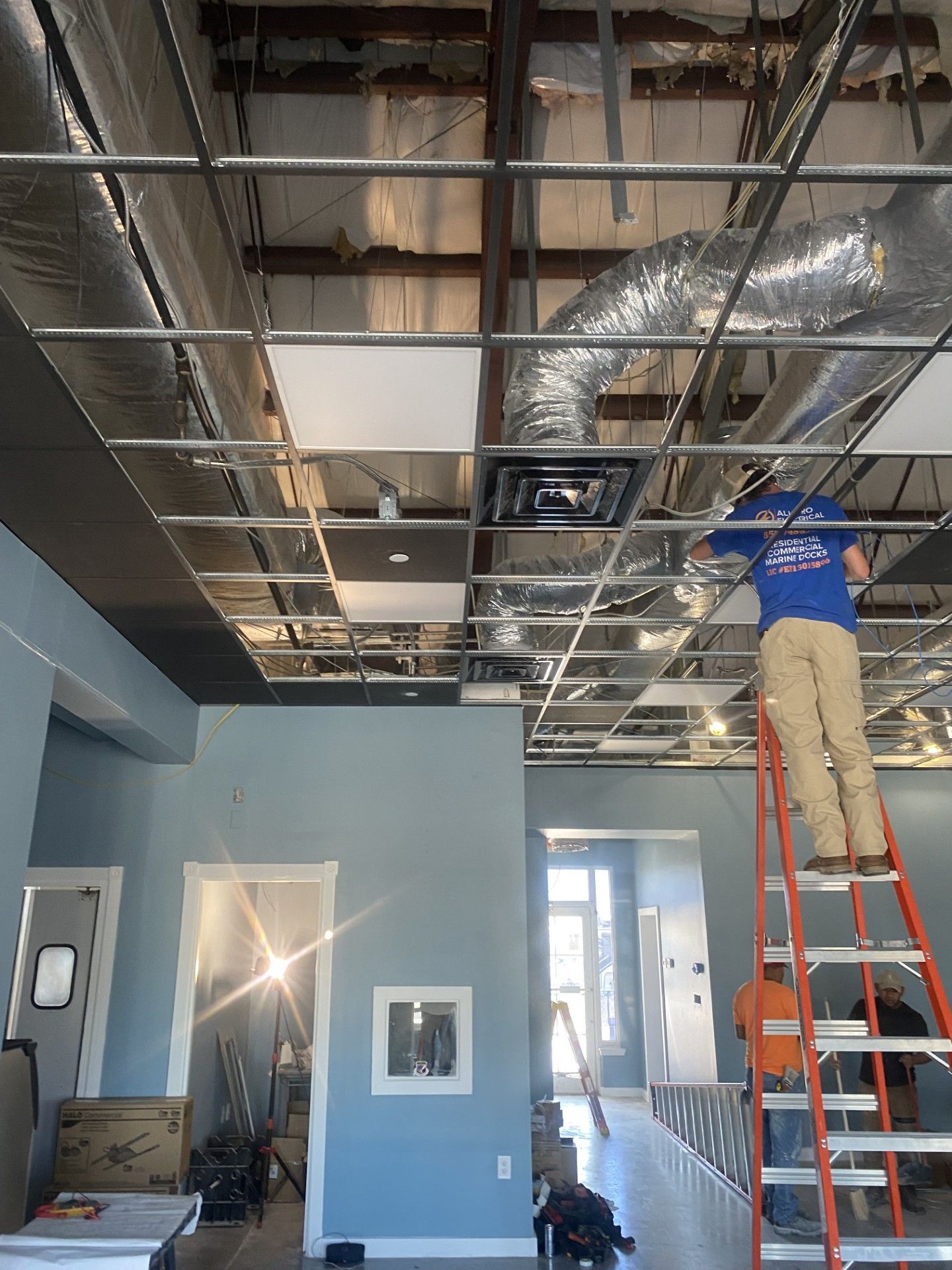 Worker on ladder installing ductwork in unfinished office ceiling.