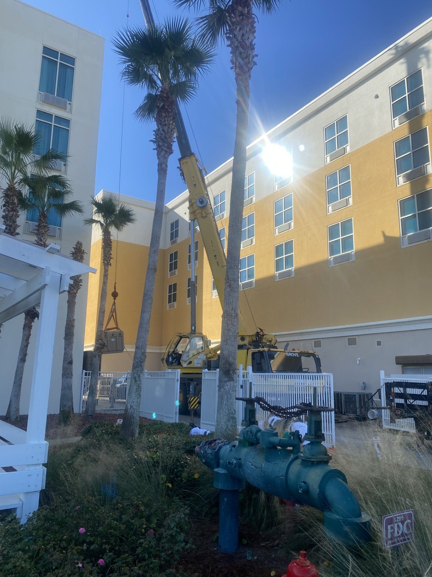 Sunlit apartment courtyard with yellow and white buildings, palm trees, and green pipes in the foreground