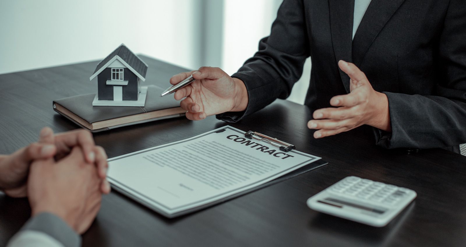 Hands exchanging a house key over a mortgage document beside a small model home and calculator