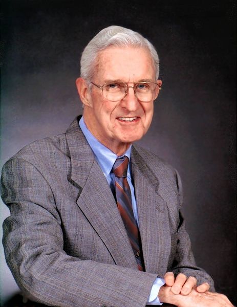 A person wearing a grey suit, blue dress shirt, and striped tie, smiling against a dark, textured background.