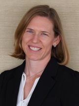 A woman with shoulder-length hair wearing a black blazer and white top, smiling against a neutral tan background.