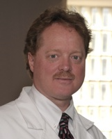 A person with short, light-brown hair and a mustache wearing a white lab coat and a patterned tie, looking at the camera.