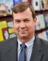 A person with short brown hair wears a gray suit and patterned tie, posing for a portrait in a library setting.