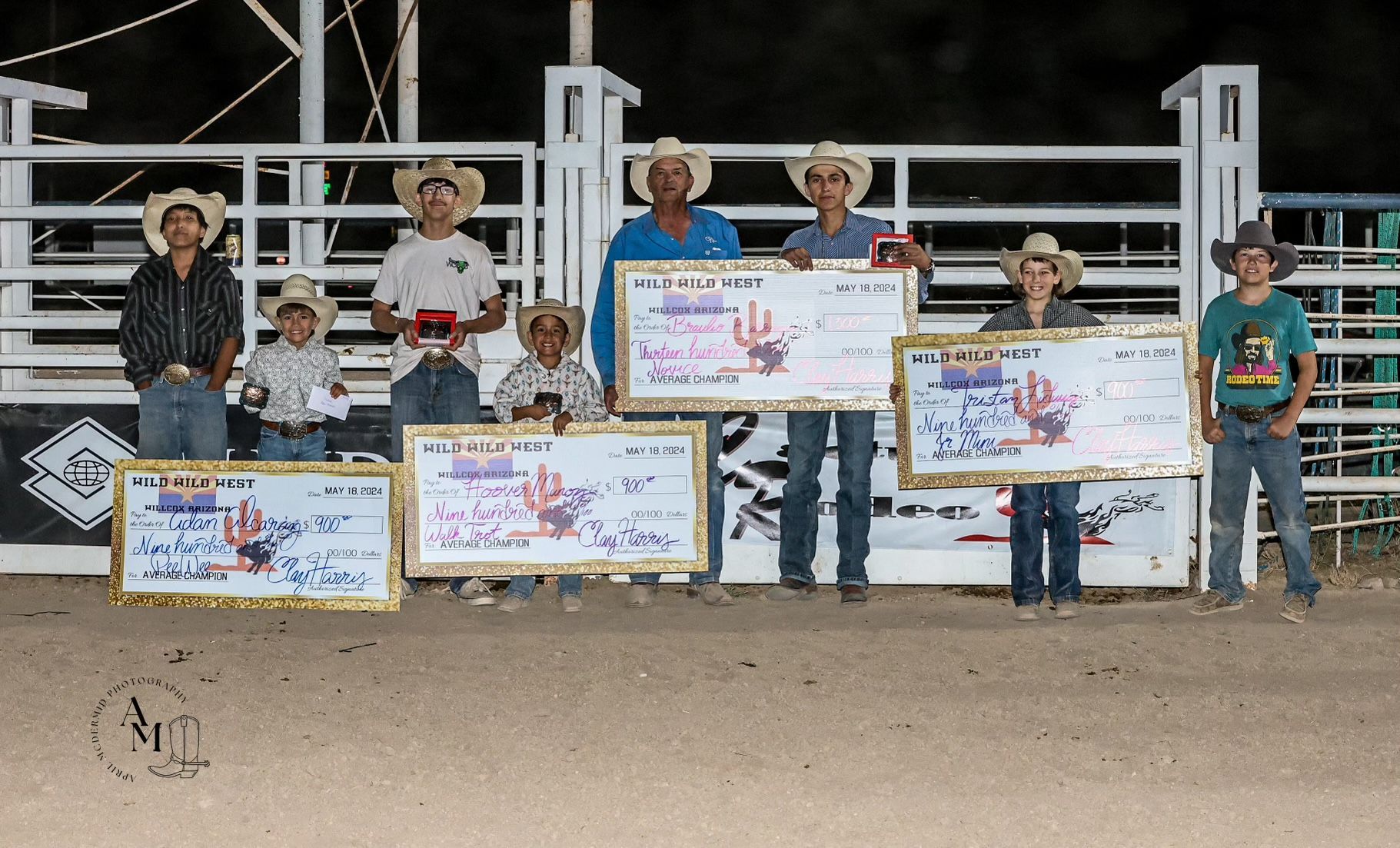 Seven youths in cowboy hats holding large checks at a rodeo, posing in front of a fence.