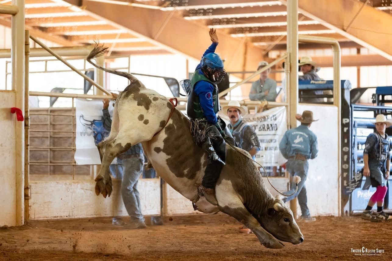 Rodeo: Bull rider airborne on bucking bull in a dirt arena, rider wearing a blue vest and helmet.