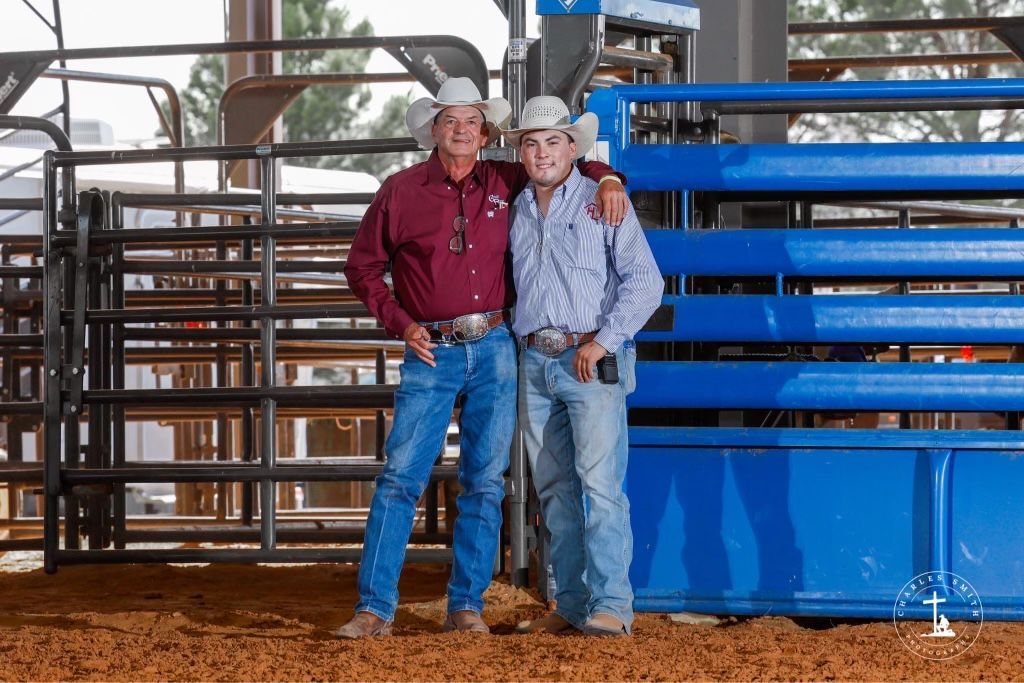 Two cowboys in hats and jeans, standing with arms around each other in a rodeo arena.