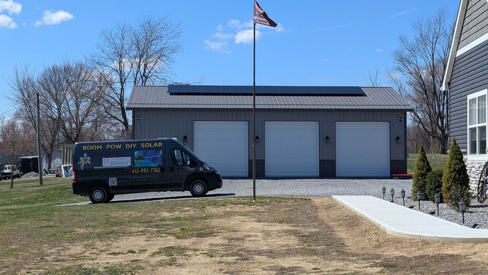 A gray van with a mural is parked in front of a three-door garage with solar panels on the roof, a flag, and a house on a sunny day.