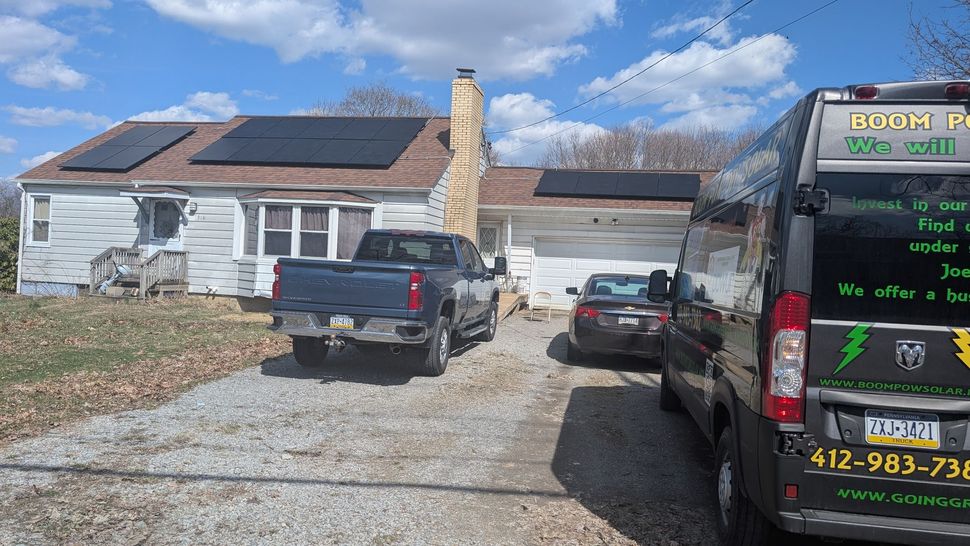 A rundown house with solar panels on the roof, a truck, a car, and a van parked in the driveway.