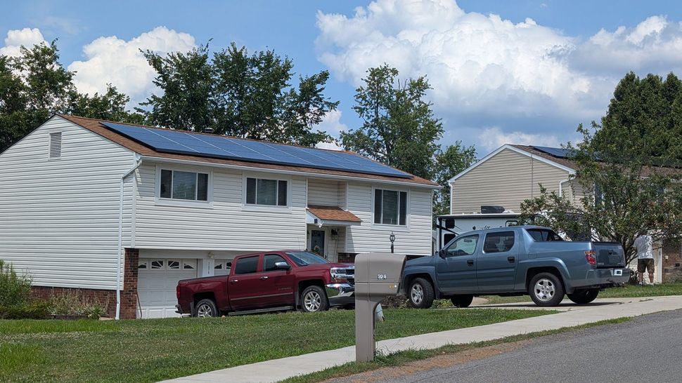 A suburban house with solar panels on its roof, a red pickup truck in the driveway, and a blue pickup truck parked on the street.