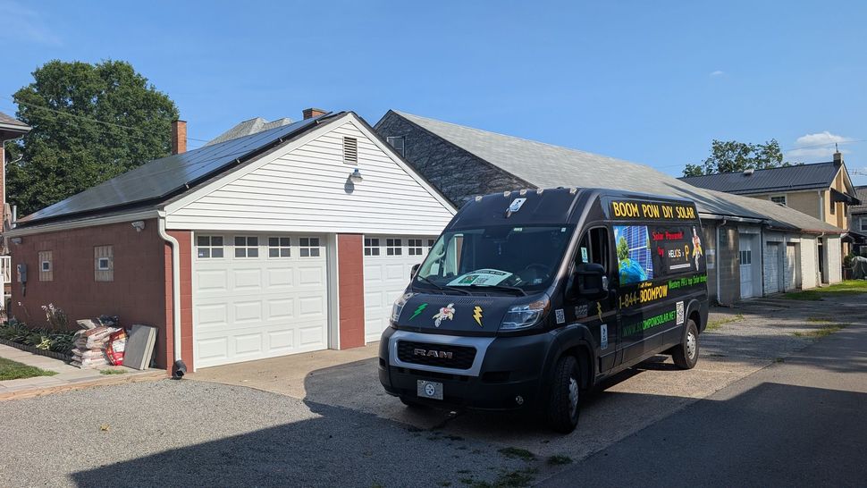 A black van with colorful logos parked in front of a garage with solar panels. The garage is red brick with a white door.