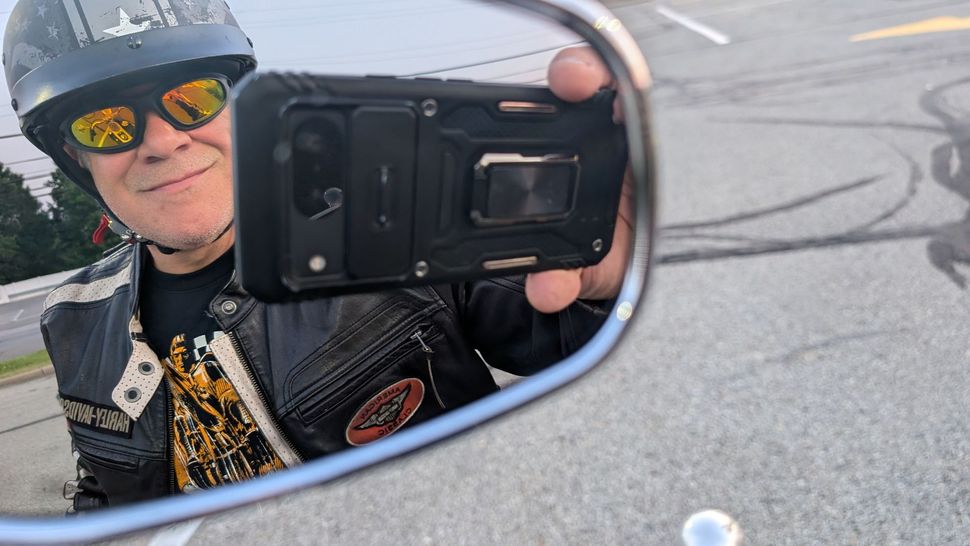 Man in motorcycle gear taking a selfie in side mirror; smiling.