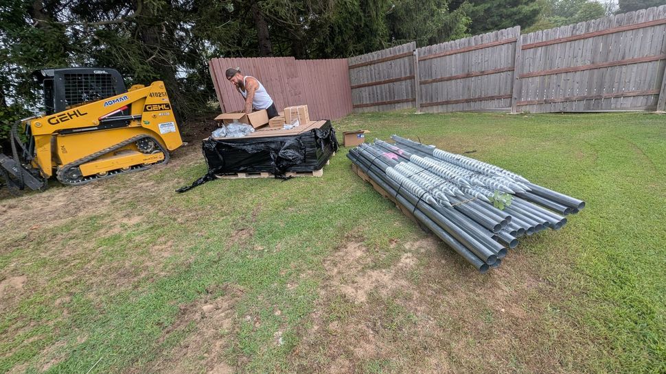 Person unloading materials next to a skid steer and fence on a grassy lawn.
