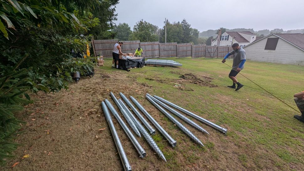 Construction workers on a grassy area; metal poles in foreground; cloudy sky.