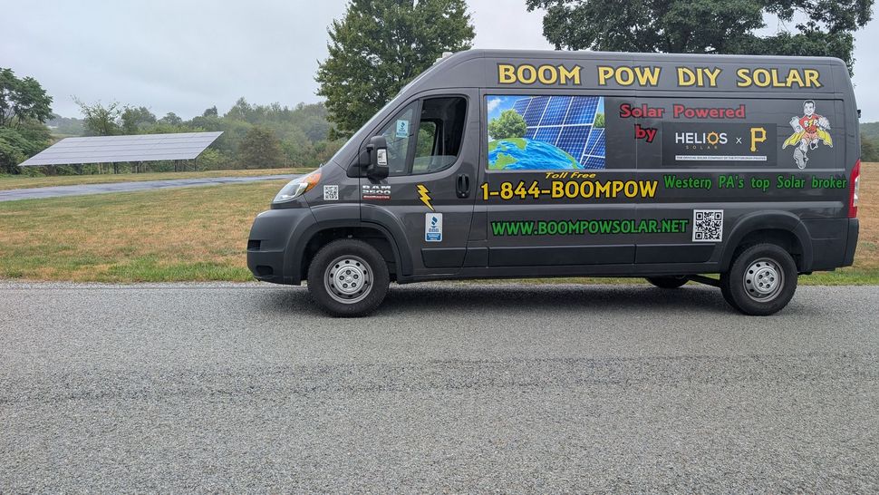 Dark van with BOOM WOW DIY SOLAR logo, parked on gravel road, solar panel field in the background.