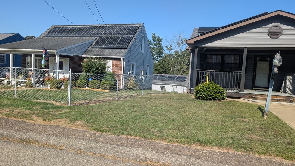 Two houses with chain-link fences in front. One has solar panels on its roof. Both have small lawns and bushes.