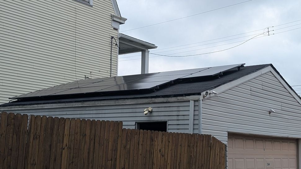 Damaged black shingle roof with missing sections on a white house behind a wooden fence