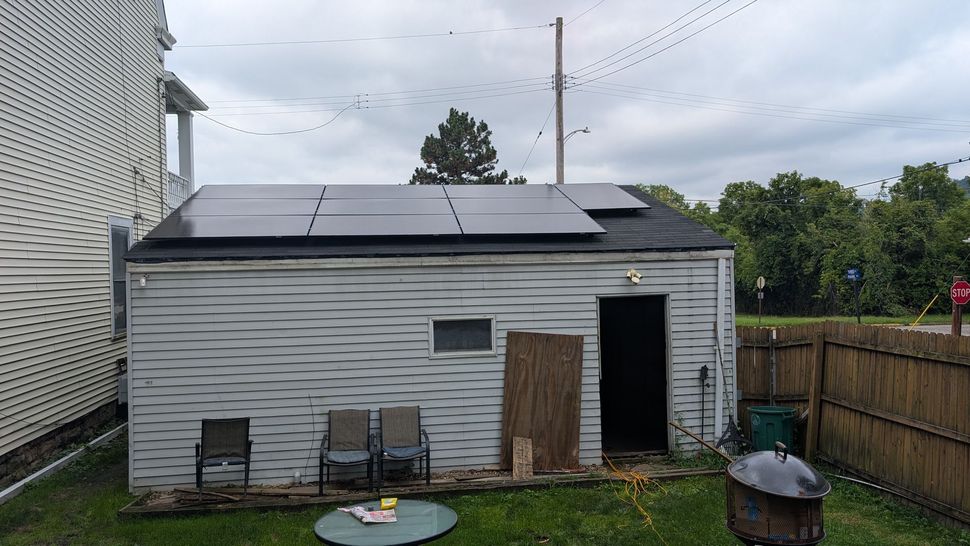 Small gray shed with solar panels in a backyard, with a fenced lawn and cloudy sky.