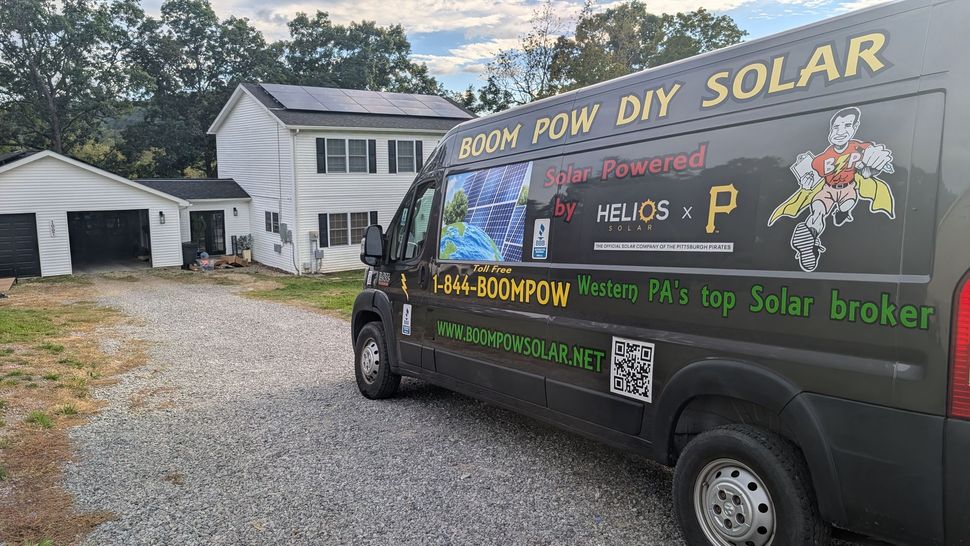 Black solar installation van parked in a driveway beside a white house and garage.