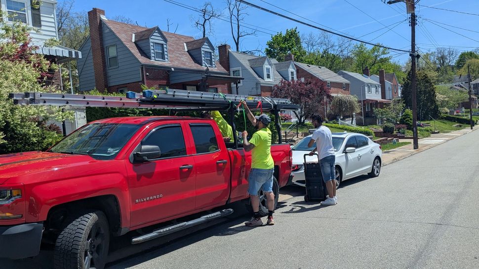 People loading a ladder onto a red truck parked on a street in front of houses on a sunny day. Two people standing next to a white car.