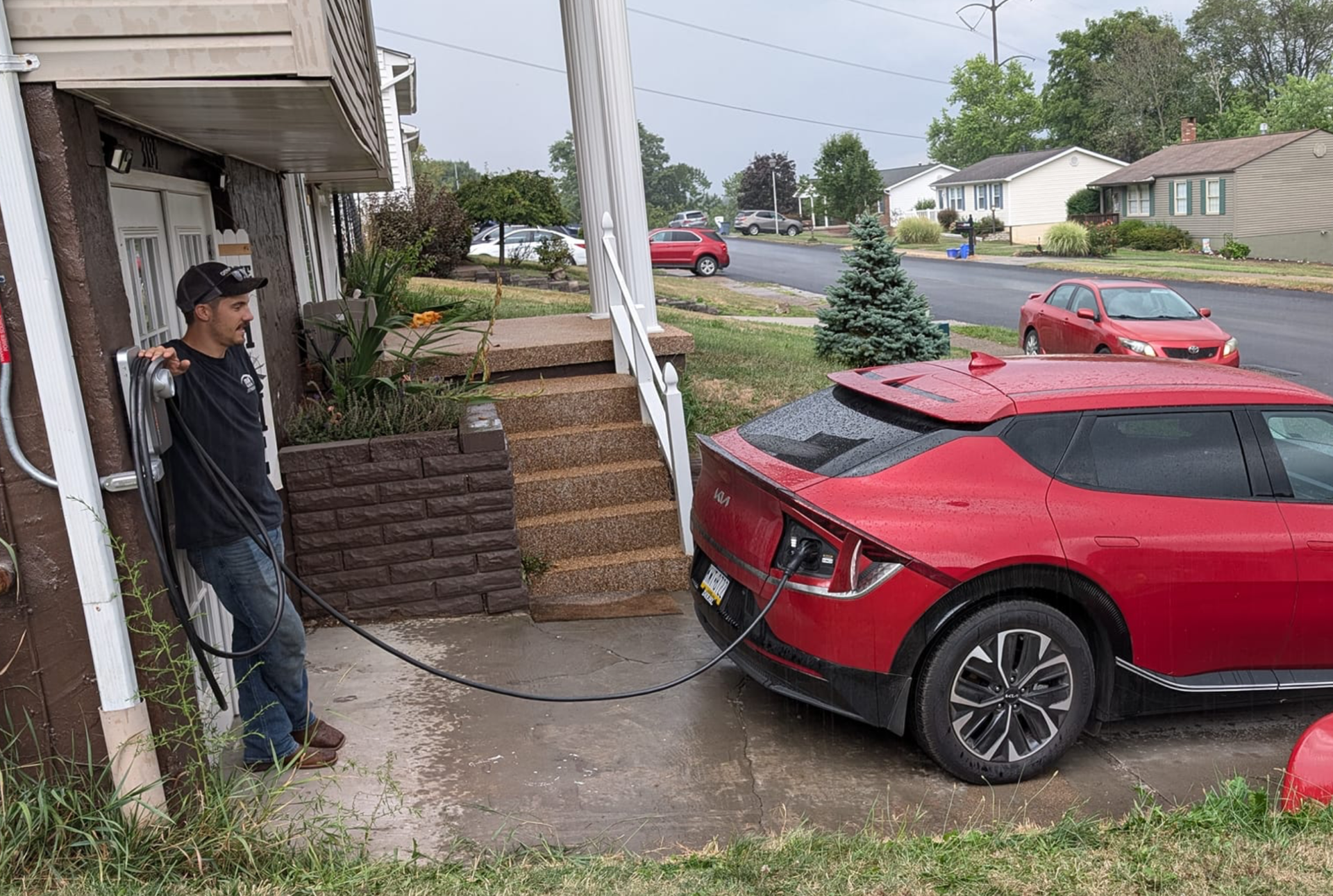 A man leans against a house while an electric car charges on the sidewalk. The red car is parked on a residential street.