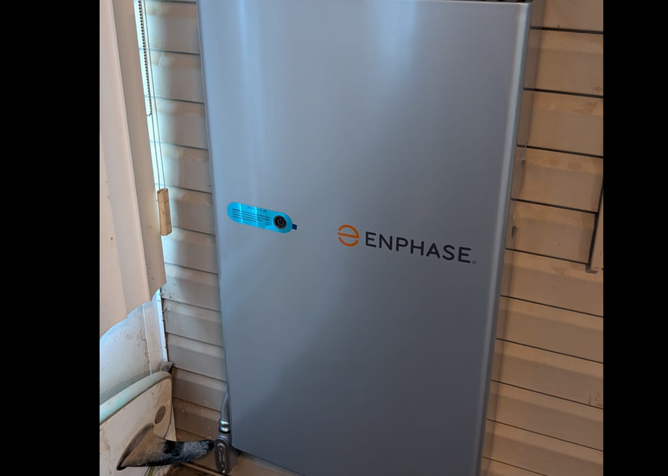 A gray Enphase battery storage unit is mounted on a wall, next to a window with white blinds.