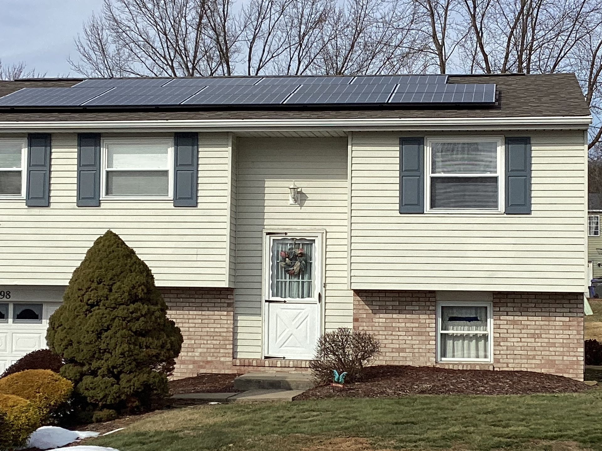 House with solar panels on roof, shutters, brick facade, and front door with wreath.
