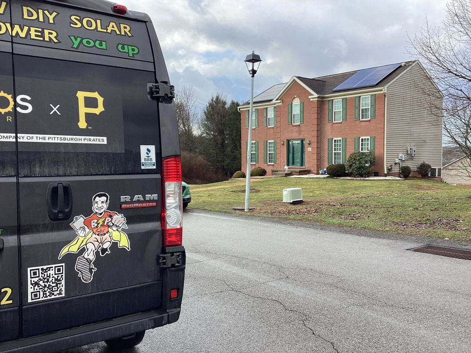 Black van with solar-power graphics parked on a suburban street beside brick houses and a lamppost