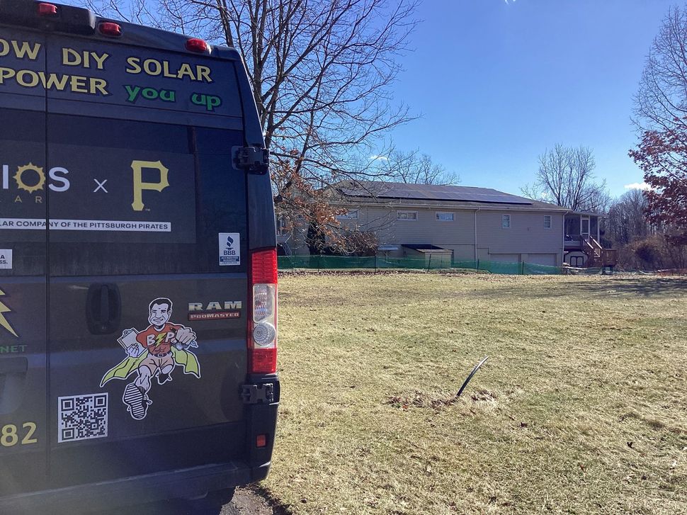 Black van with solar power graphics parked on grassy field, house in background.