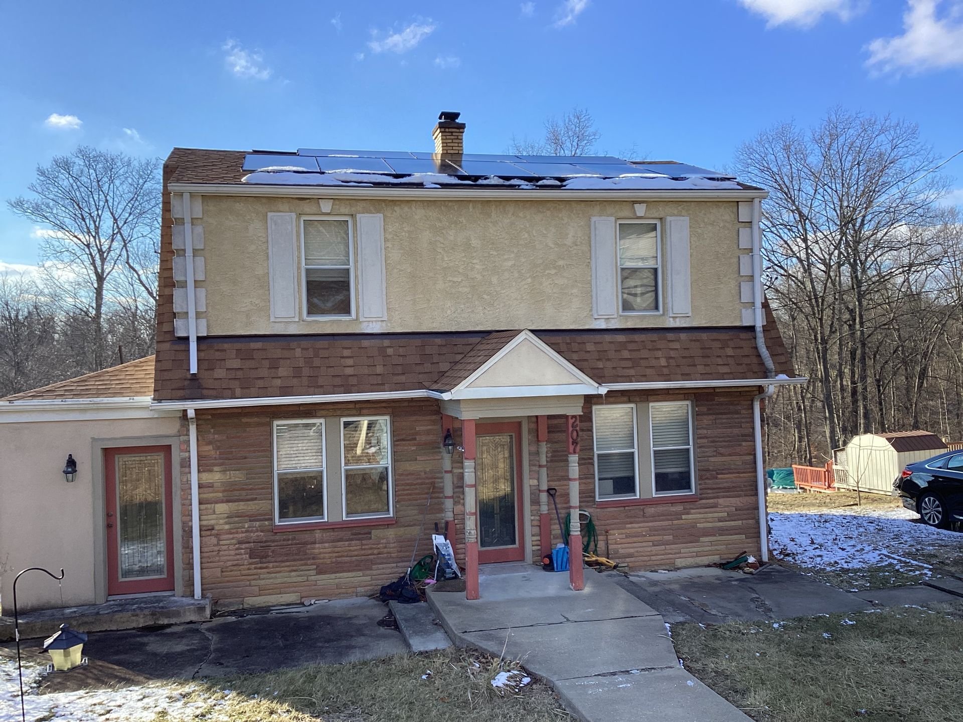 Two-story brick and stucco house with snow on roof, porch, and bare trees in the background.