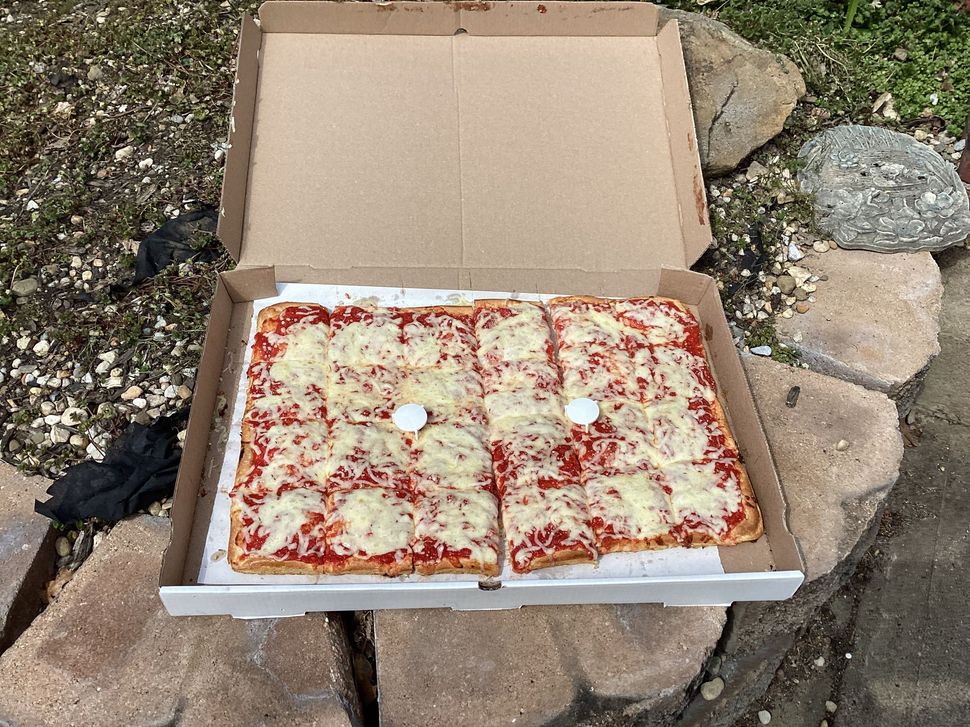 A square-cut cheese pizza in an open cardboard box, resting on a stone surface outdoors.