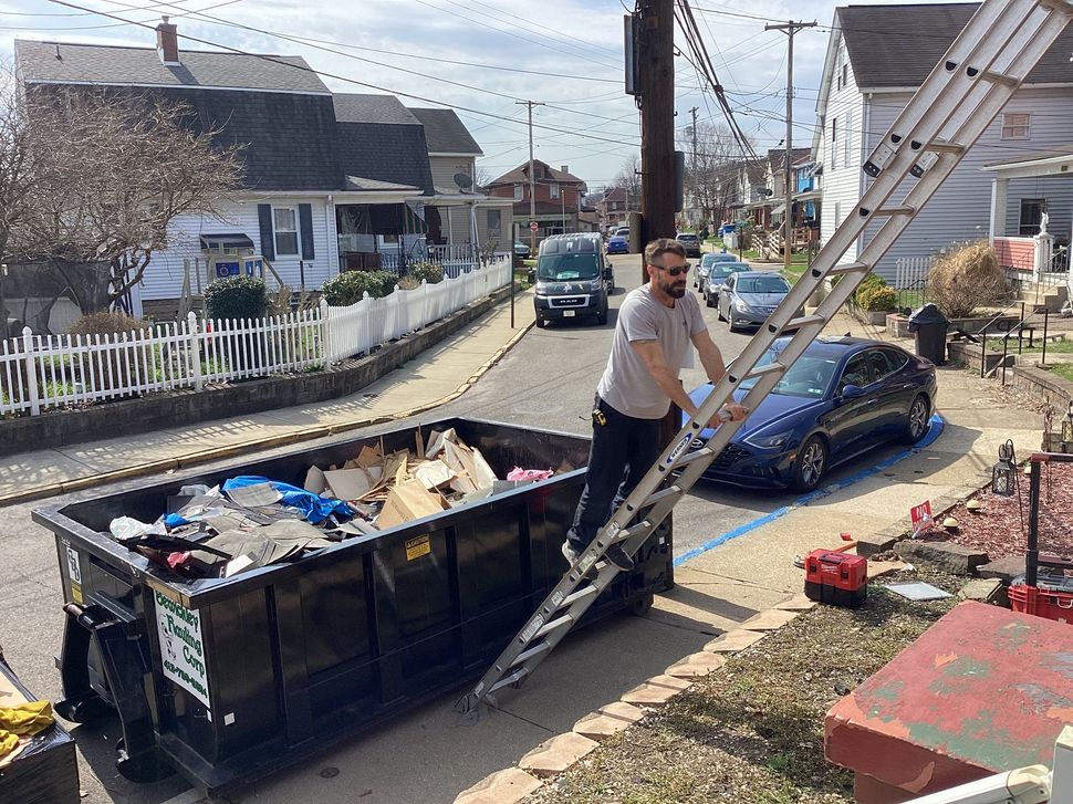 Man on a ladder beside a dumpster and blue car near a damaged curbside scene