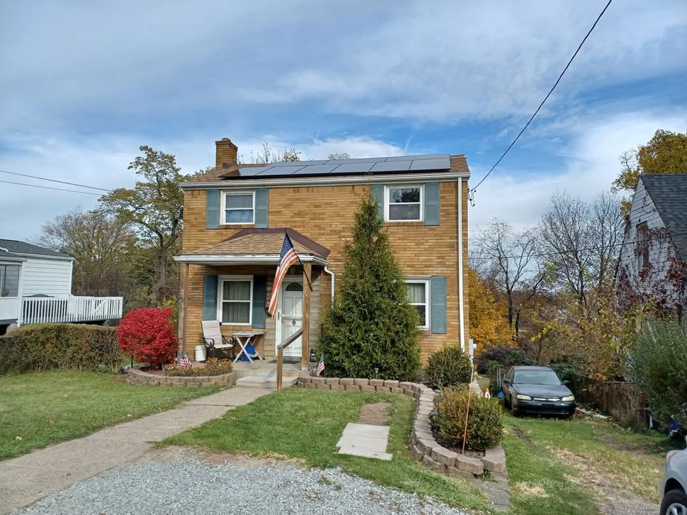 Two-story brick house with green shutters, solar panels, and a small front yard; a car is parked on the right.