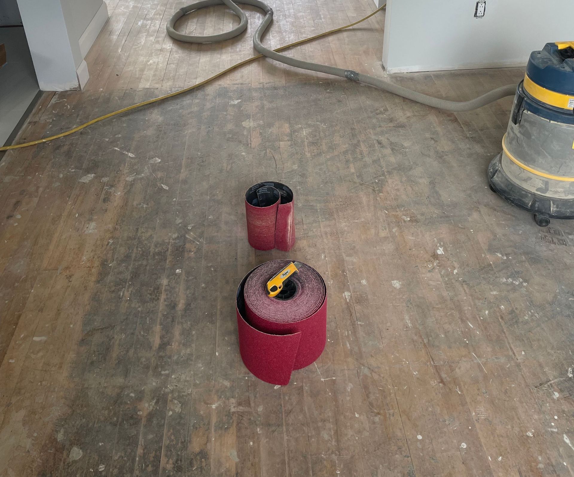 Bandes abrasives rouges sur un plancher en bois poussiéreux, près d'un aspirateur et d'un tuyau.