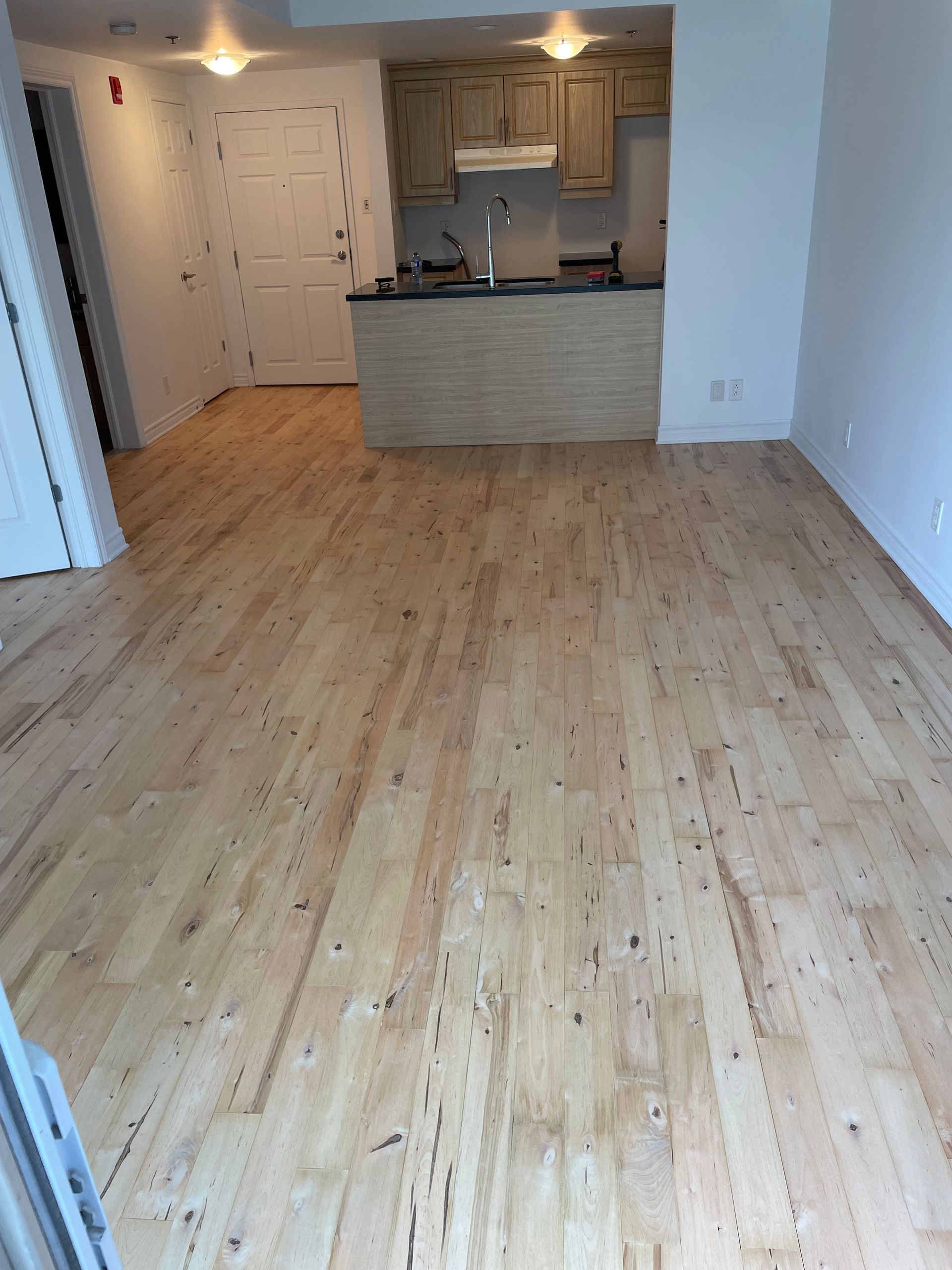 Sol en bois clair dans une pièce, avec une cuisine visible en arrière-plan. On y distingue des placards et un plan de travail.