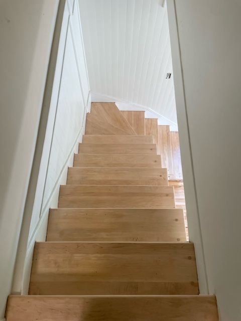 Escalier en bois, vu d'en bas, menant vers le haut, entre des murs blancs, jusqu'à un plafond blanc.