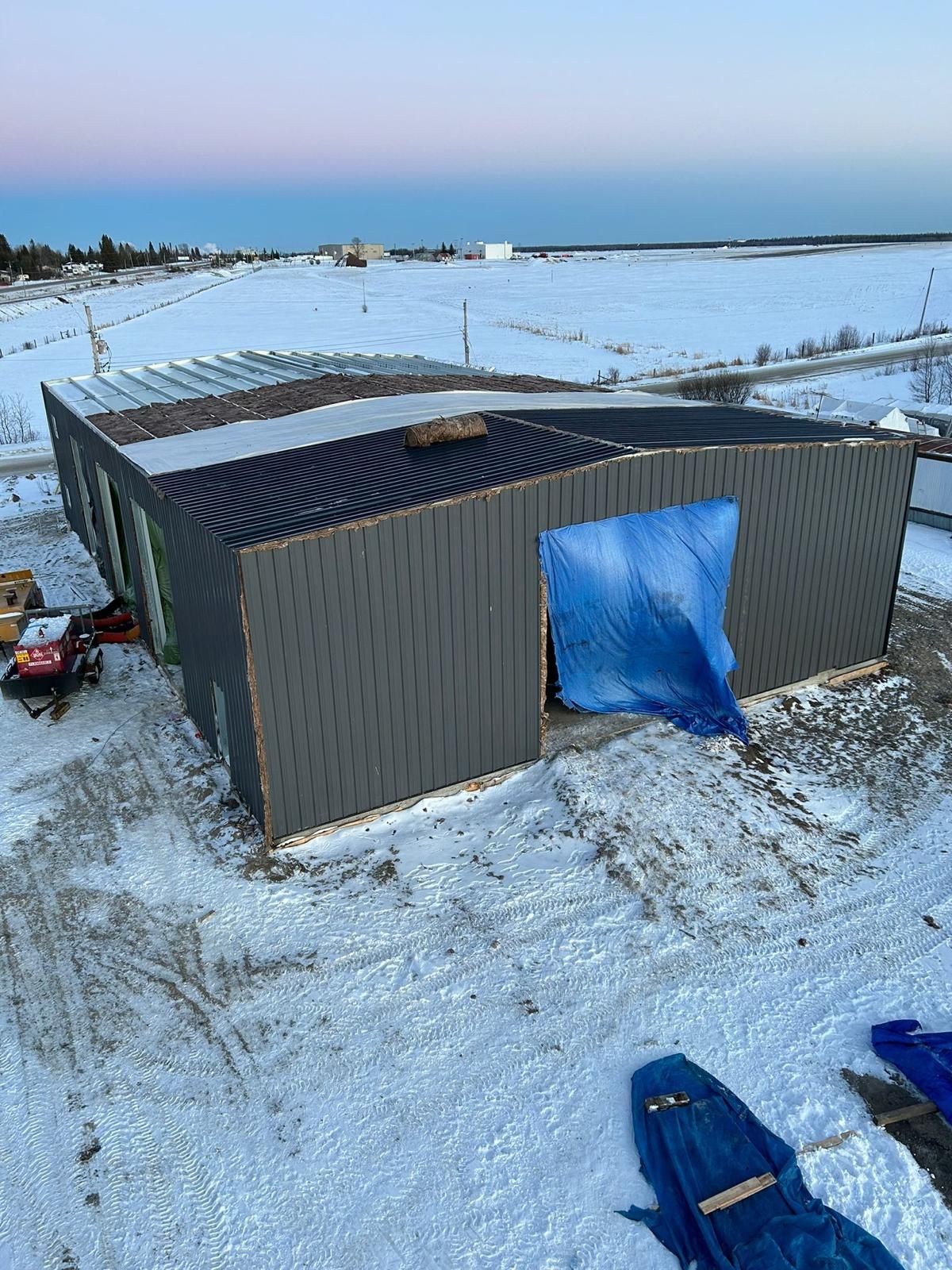 Small metal shed in a snowy field with a blue tarp-covered doorway and corrugated roof.